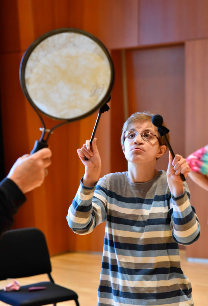 A young person plays a large frame drum held up by another person