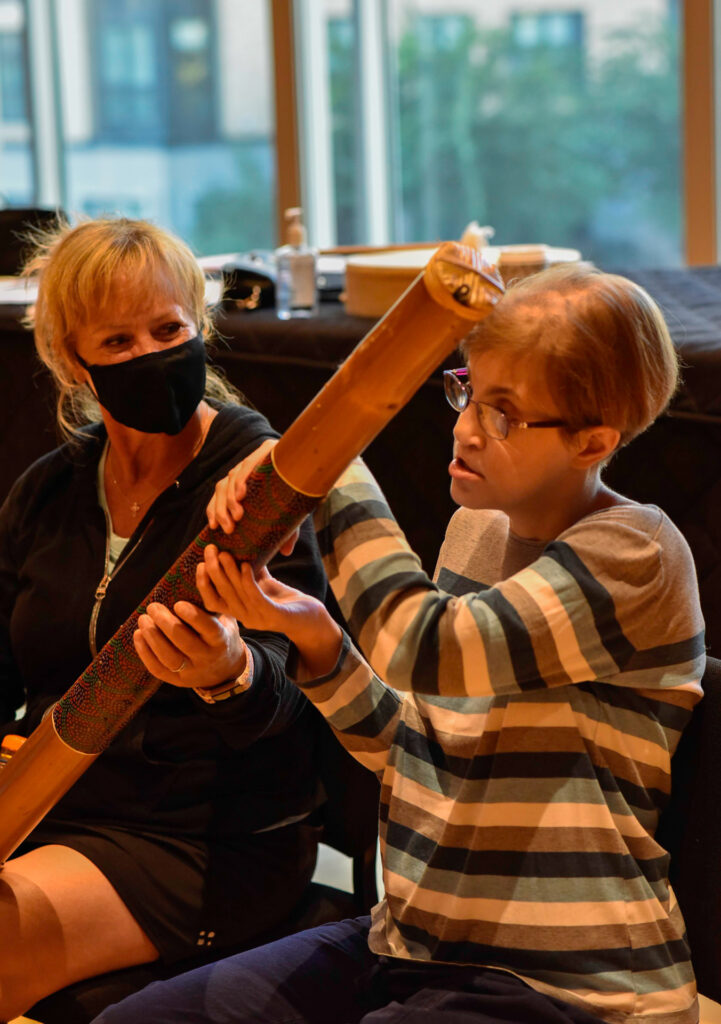 A woman helps a young person hold a long wooden percussion tube