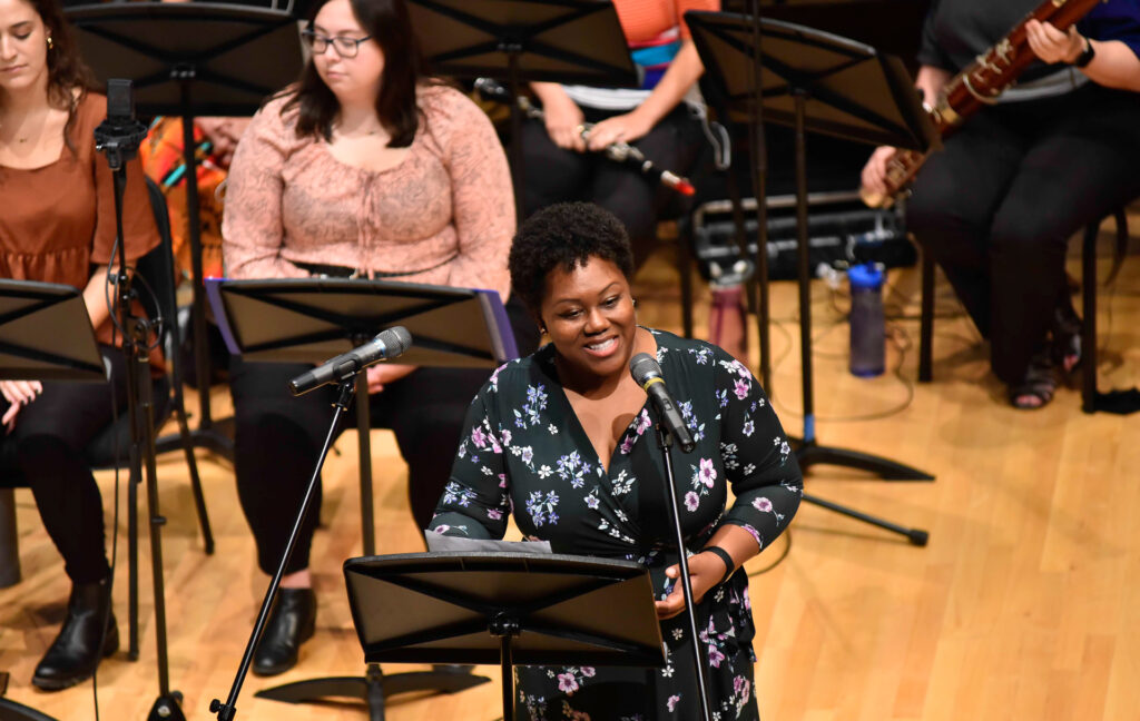 A woman in a floral dress sings into a microphone in front of the ensemble