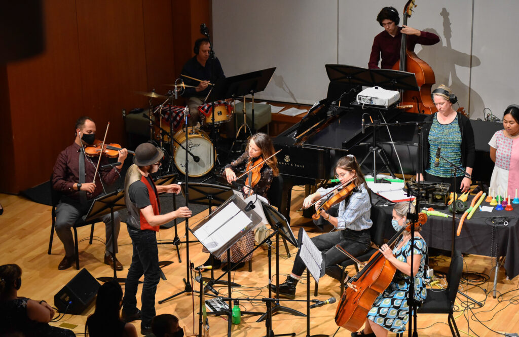 A group of musicians performs together on stage, including violinists, a cellist, a standing bassist, and percussionists at a table