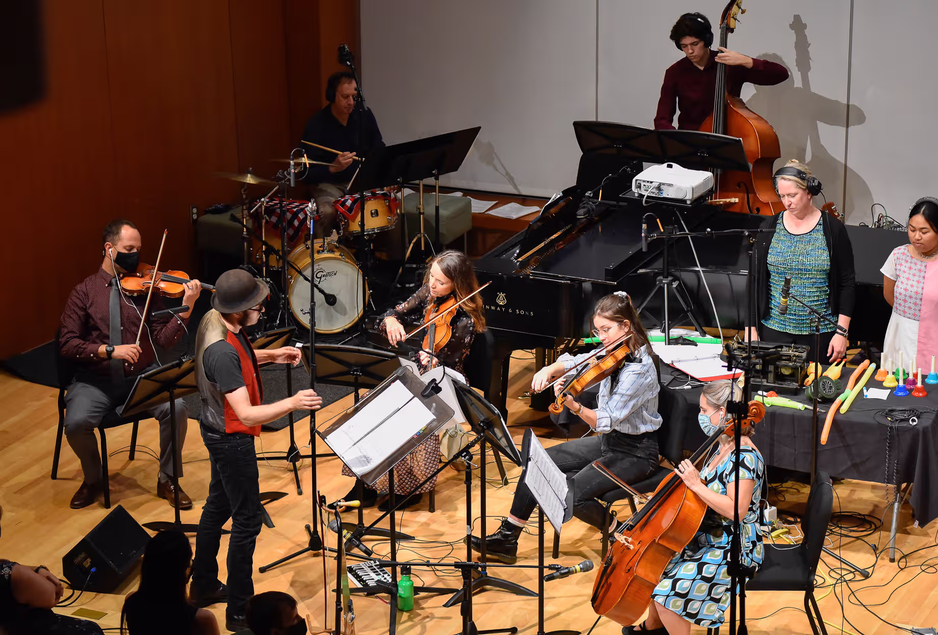 A musical ensemble with string players, a drummer, and a conductor performing on a stage with recording equipment