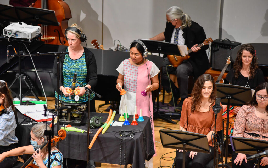 Two women wearing headphones shake percussion instruments together on stage, with a guitarist playing behind them