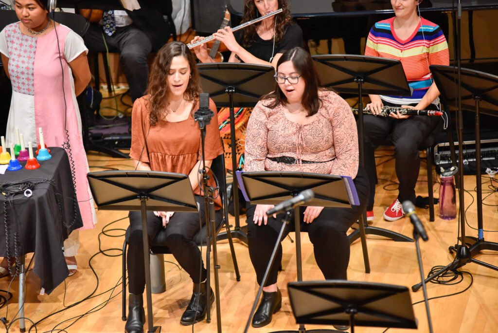 Two women sit at microphones with sheet music stands while a flutist plays behind them