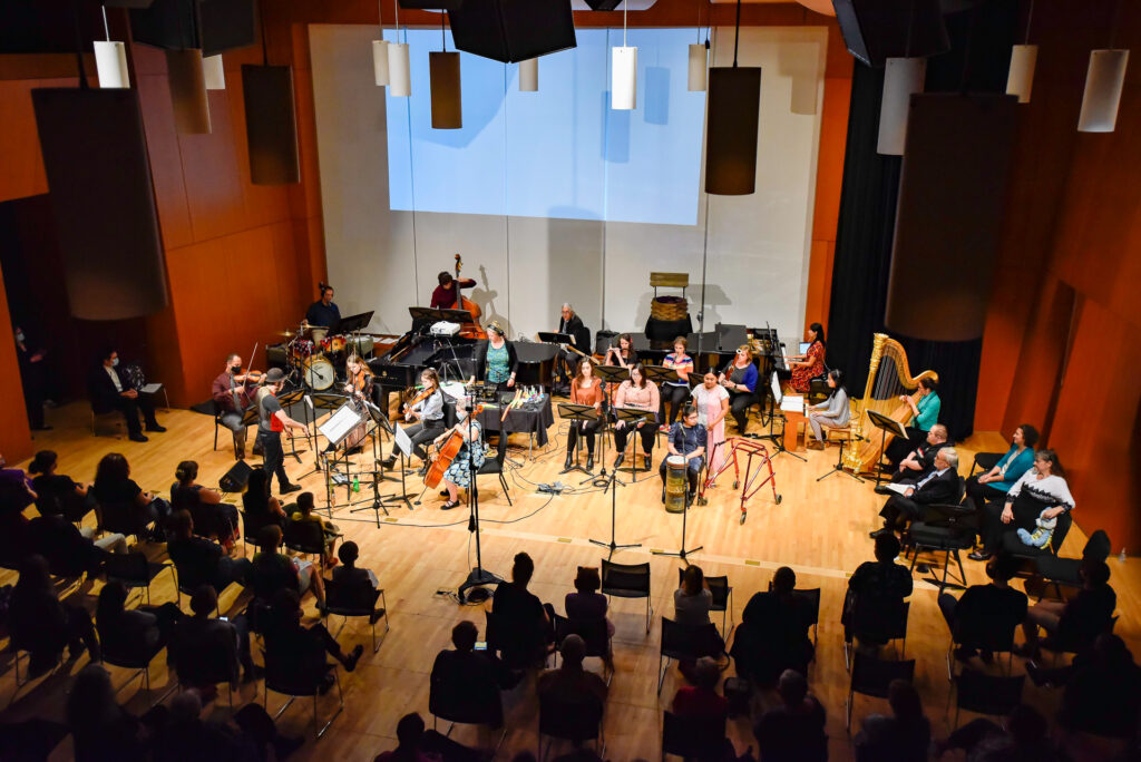 A wide view of the full orchestra performing on stage with the audience seated in front