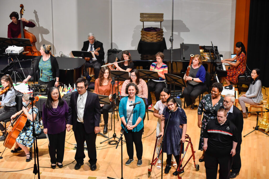 The full cast and orchestra stand together on stage, including a musician using a walker in front