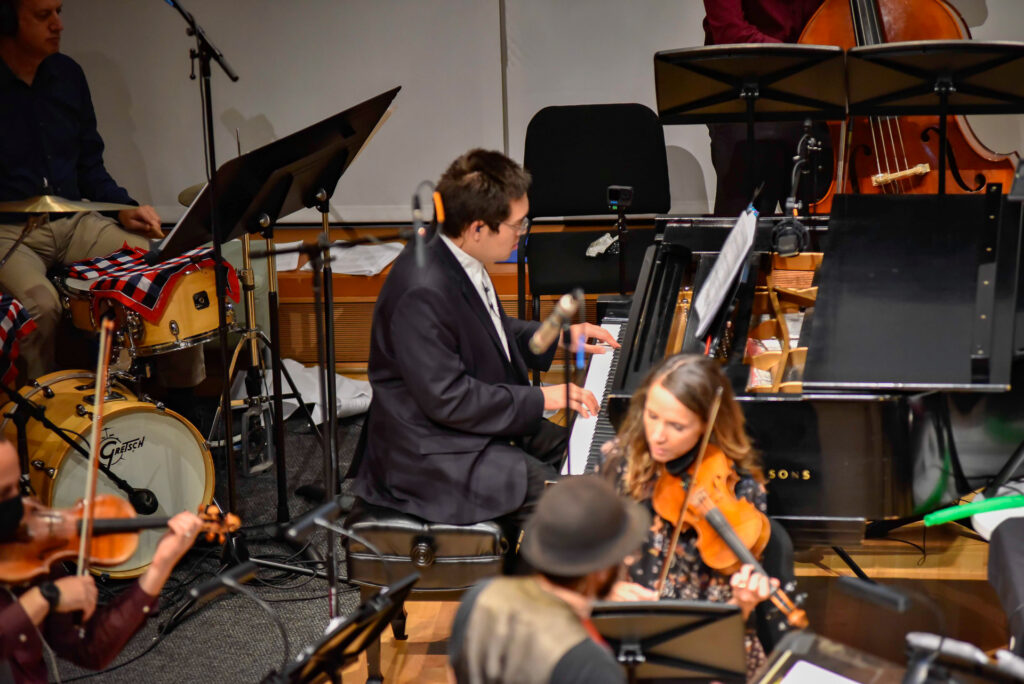 A man in a suit plays piano surrounded by other musicians, including a violinist and drummer