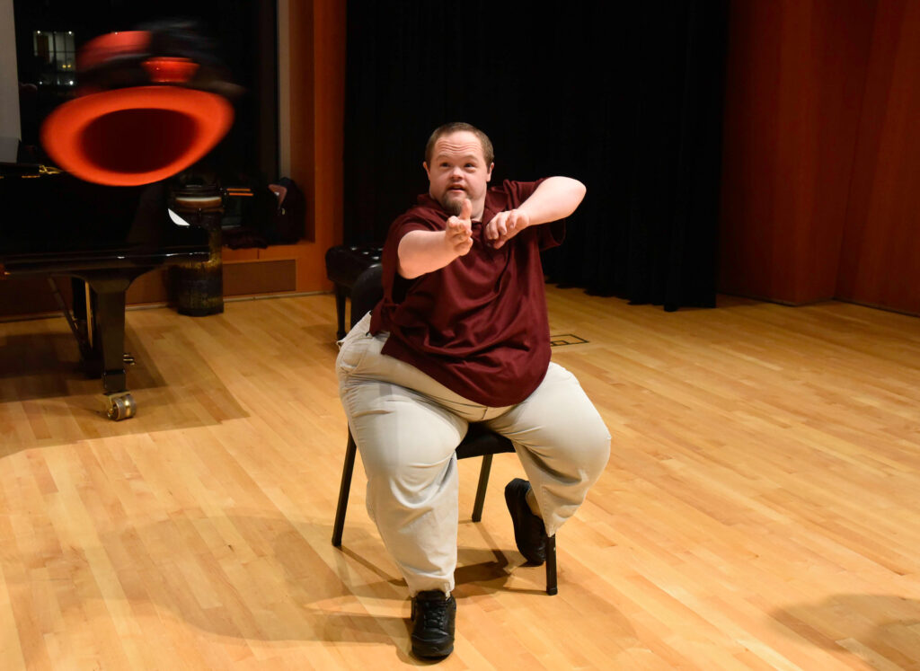 A man sits on a chair mid-motion after tossing his red top hat into the air