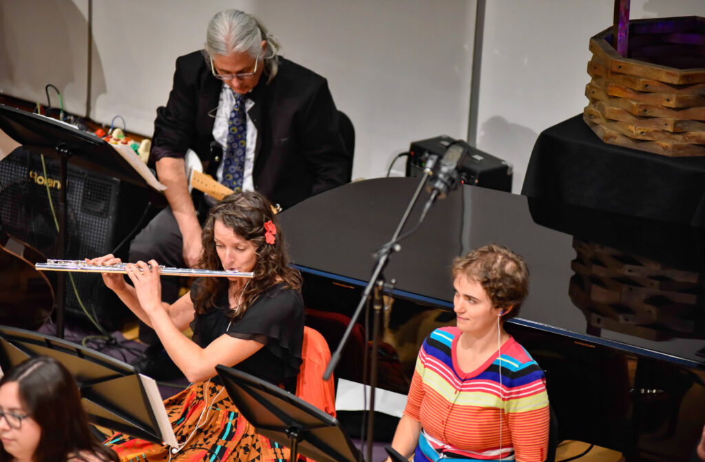 A woman plays the flute while a man plays electric guitar behind her on stage