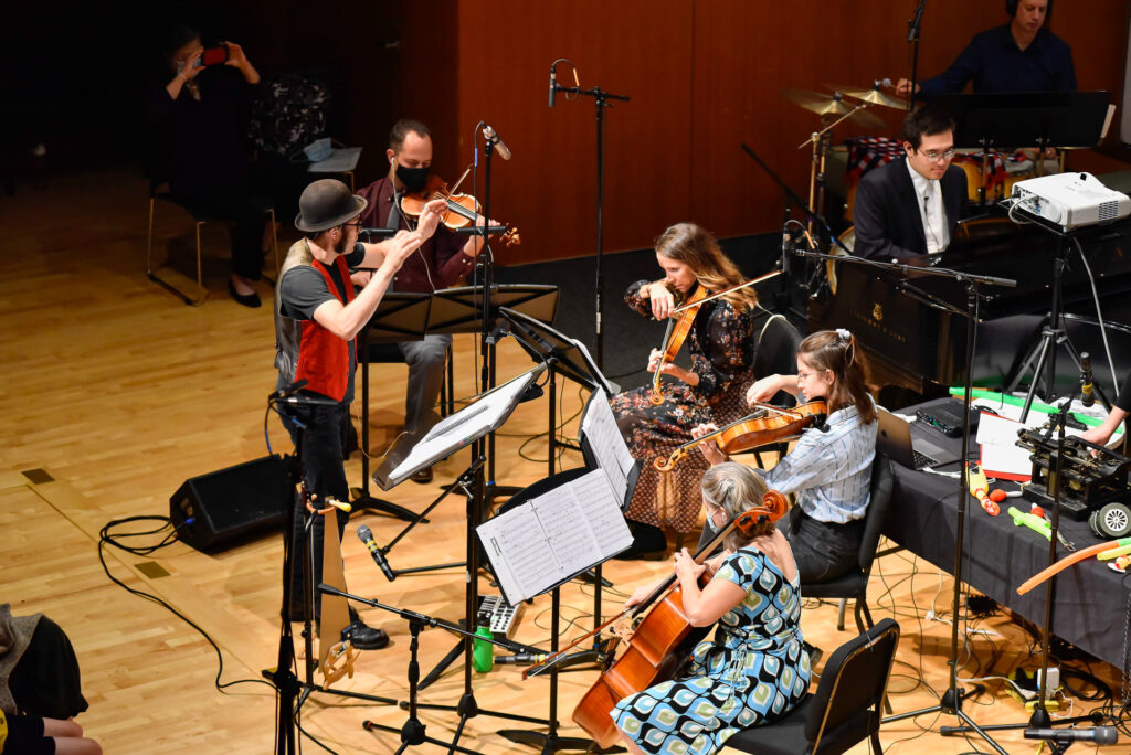 An ensemble of musicians performs on stage, including violinists, a cellist, and a pianist