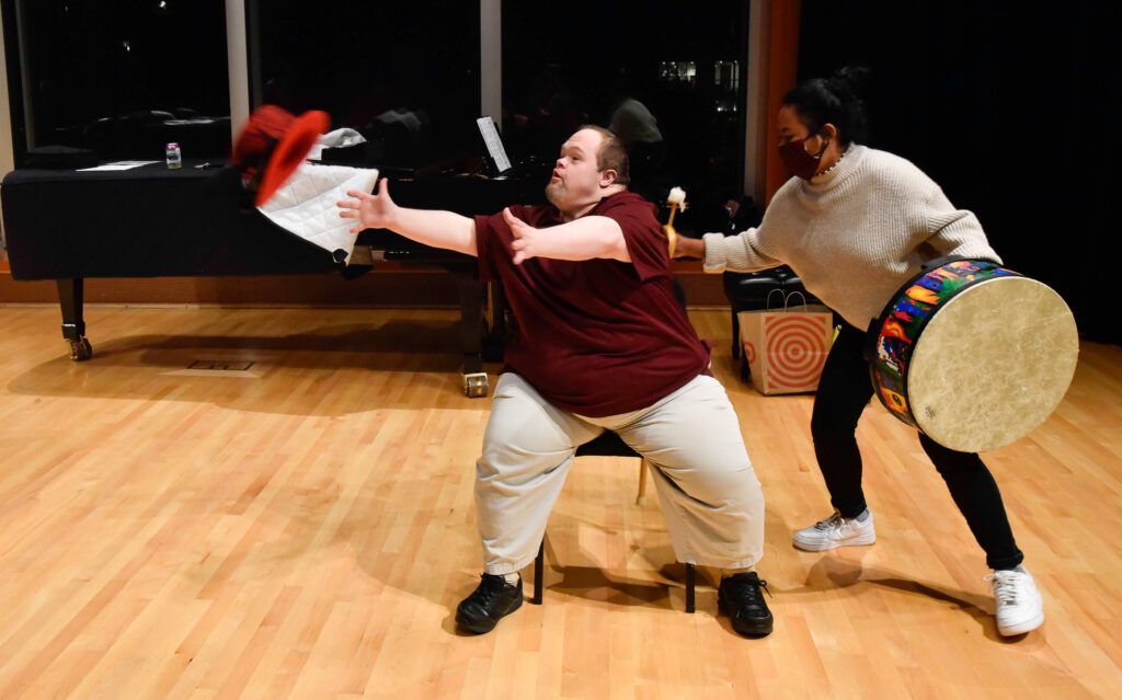 A man tosses a red top hat into the air beside a woman holding a large hand drum, with a piano visible behind them