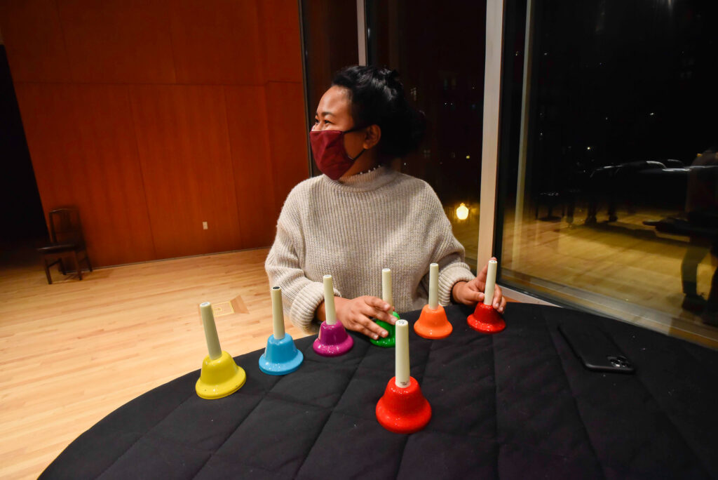 A woman wearing a mask sits at a table with a row of colorful handbells