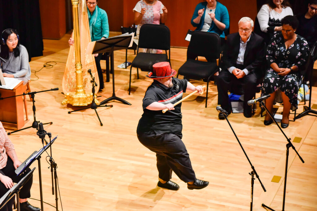 A man wearing a red top hat dances on stage with a cane while seated musicians and audience members watch