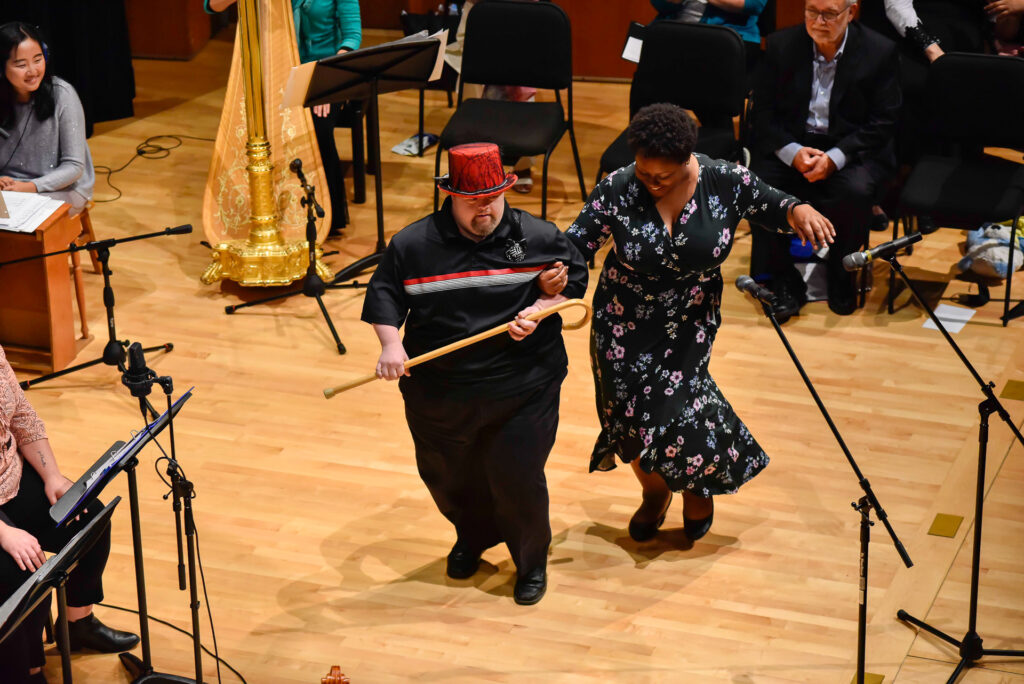 A man wearing a red top hat holds a cane while walking across the stage with a woman, musicians seated behind them