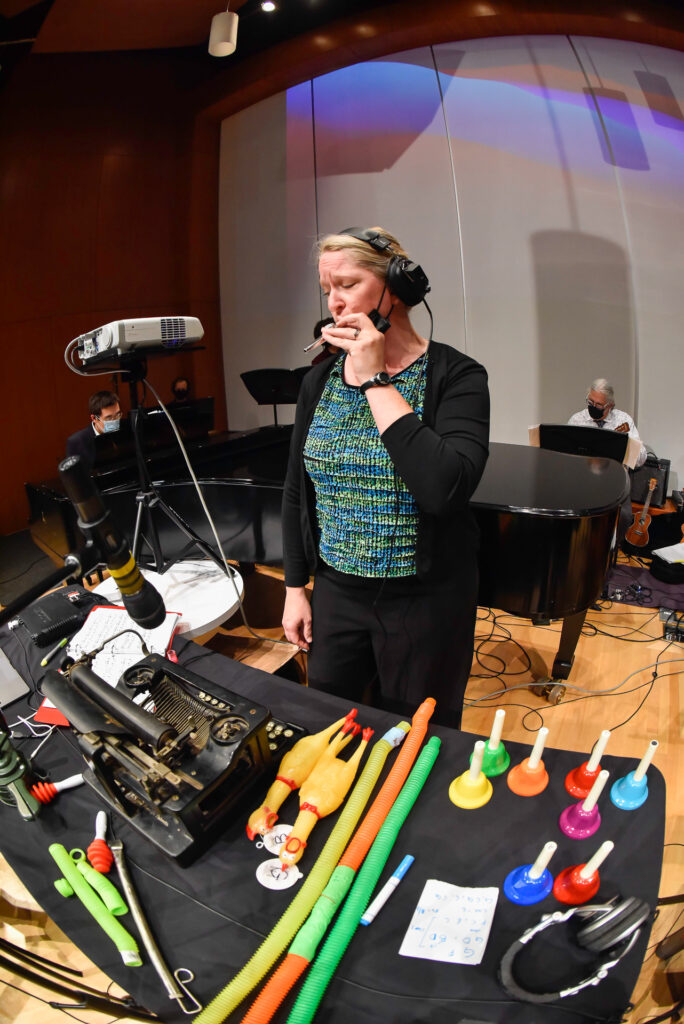 A woman wearing headphones speaks into a microphone beside a table of colorful percussion instruments, with a pianist visible on stage behind her