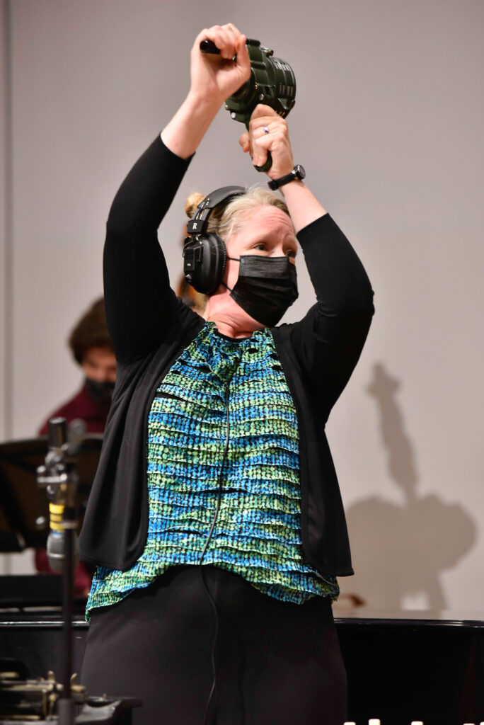 A woman wearing headphones and a mask raises a percussion shaker instrument overhead