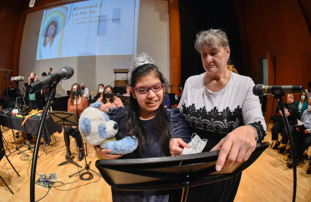 A girl wearing a tiara holds a stuffed dog toy at a microphone stand beside a woman, with a screen behind them reading "Movement 2: Let Me Try"