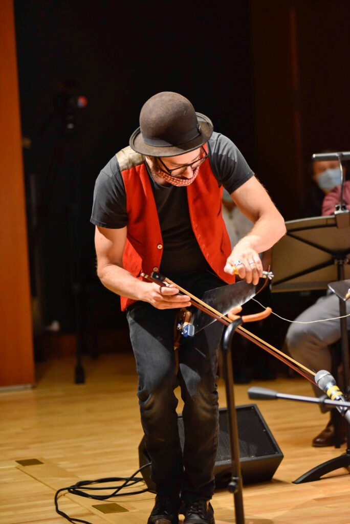 A man wearing a bowler hat and red vest plays a handmade stringed instrument with a bow