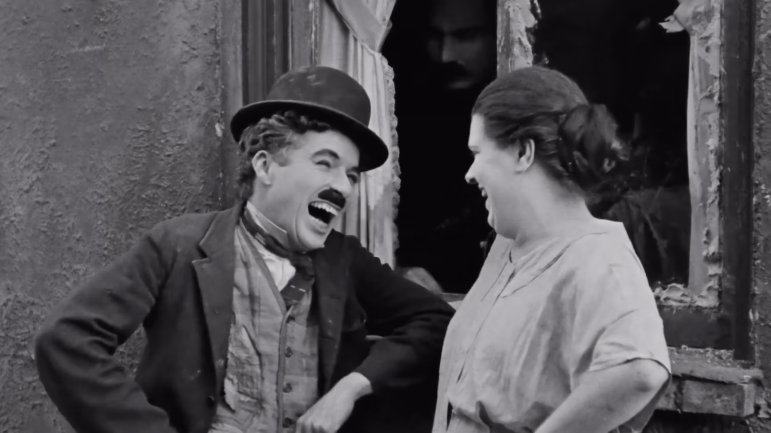 Charlie Chaplin as the Tramp and a woman laugh together outside a window while a man watches from the shadows