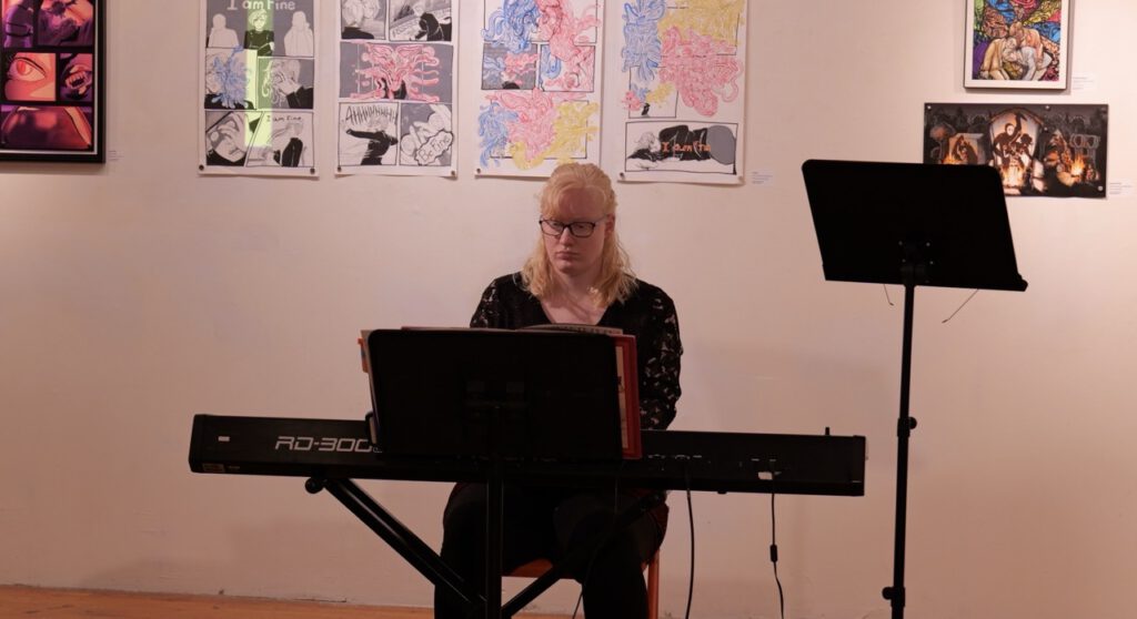 A musician plays an electric keyboard during a performance in an art gallery with artwork displayed on the wall behind her.