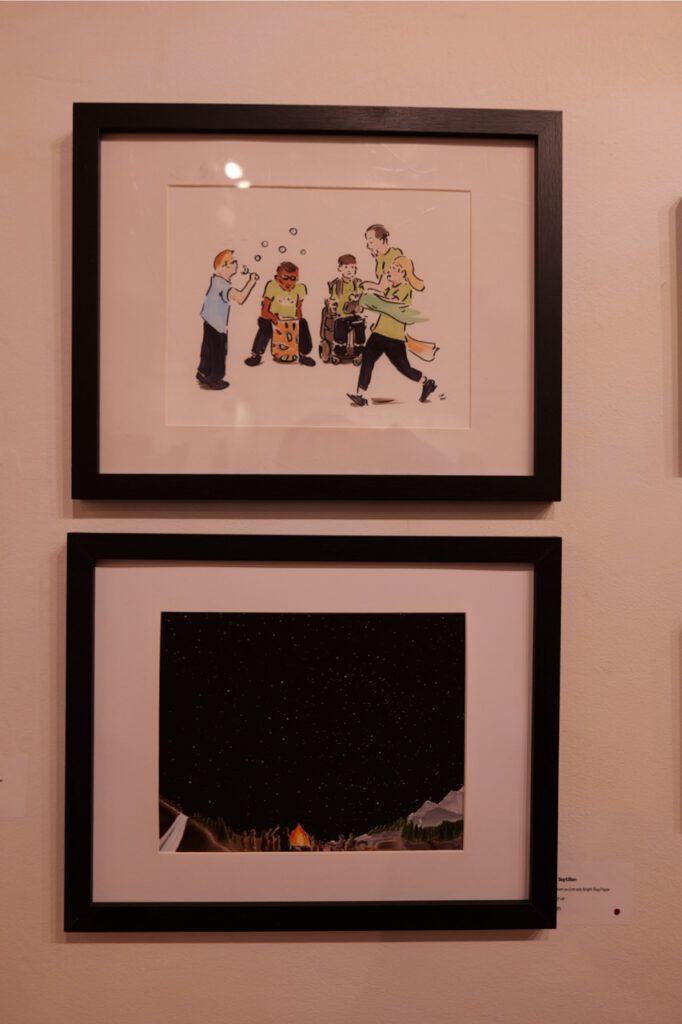 Two black-framed artworks hanging on a gallery wall: top frame depicts children playing drums and blowing bubbles; bottom frame shows a starry night sky over a campfire.