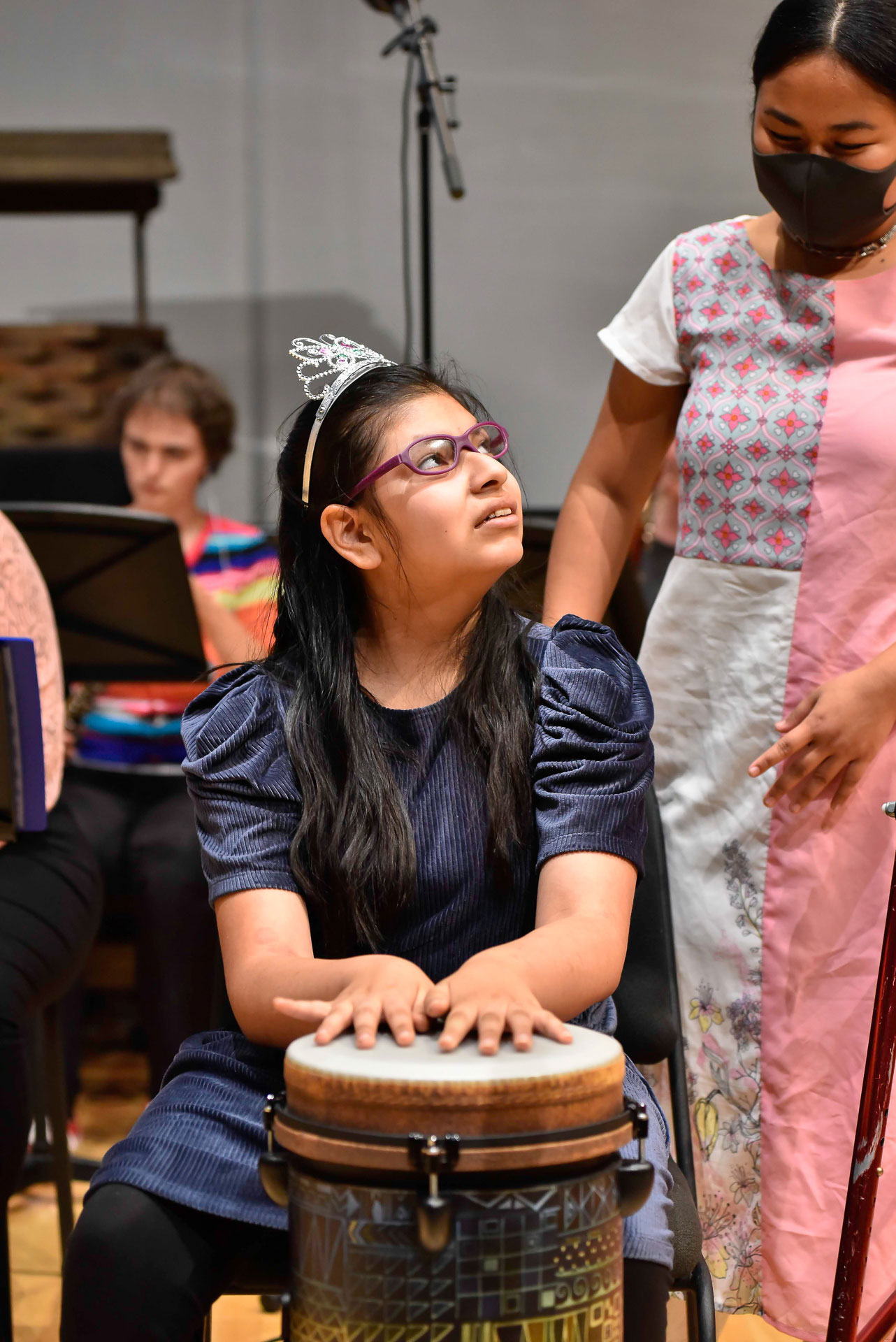 A girl wearing a tiara plays a hand drum on stage while an assistant stands behind her