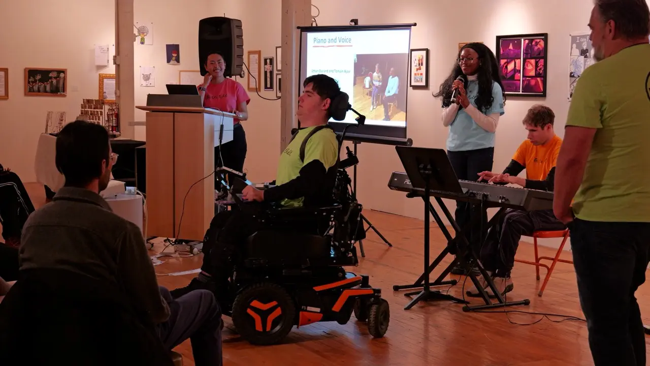 A man in a power wheelchair, a woman with a microphone, and a man playing a keyboard perform in an art gallery