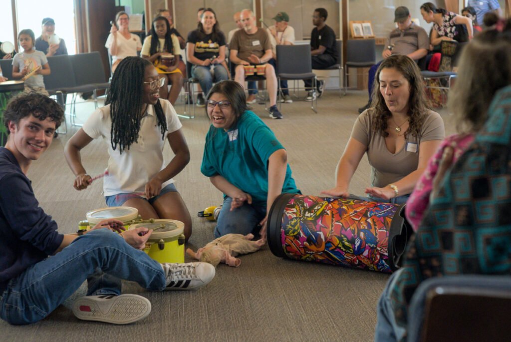 A diverse group sitting on the floor laughs and plays hand drums together in a circle
