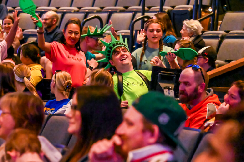 Audience members wearing Statue of Liberty crowns and Be the Light shirts cheering in a theater auditorium