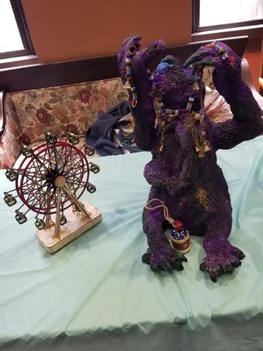Table-top sculpture of a purple dragon figure sitting upright with arms raised, strung with beads and small bells, beside a miniature red ferris wheel model